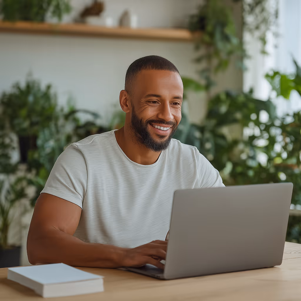 Smiling man with a beard working on a laptop at a wooden table with green plants in the background.